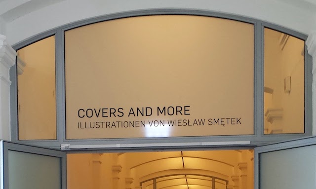 A glass window above the open door with text "COVERS AND MORE. ILLUSTRATIONEN VON WIESŁAW SMĘTEK."