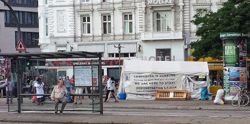 A picture of a big white tent in front of a Molike Hotel. The tent carries a sign "LAMPEDUSA IN HAMBURG VICTIMS OF NATO WAR 2011 IN LYBIA WE ARE HERE TO STAY! DEMONSTRATION 5.7.2014 12:00h NACHMANPLATZ" [original spelling]